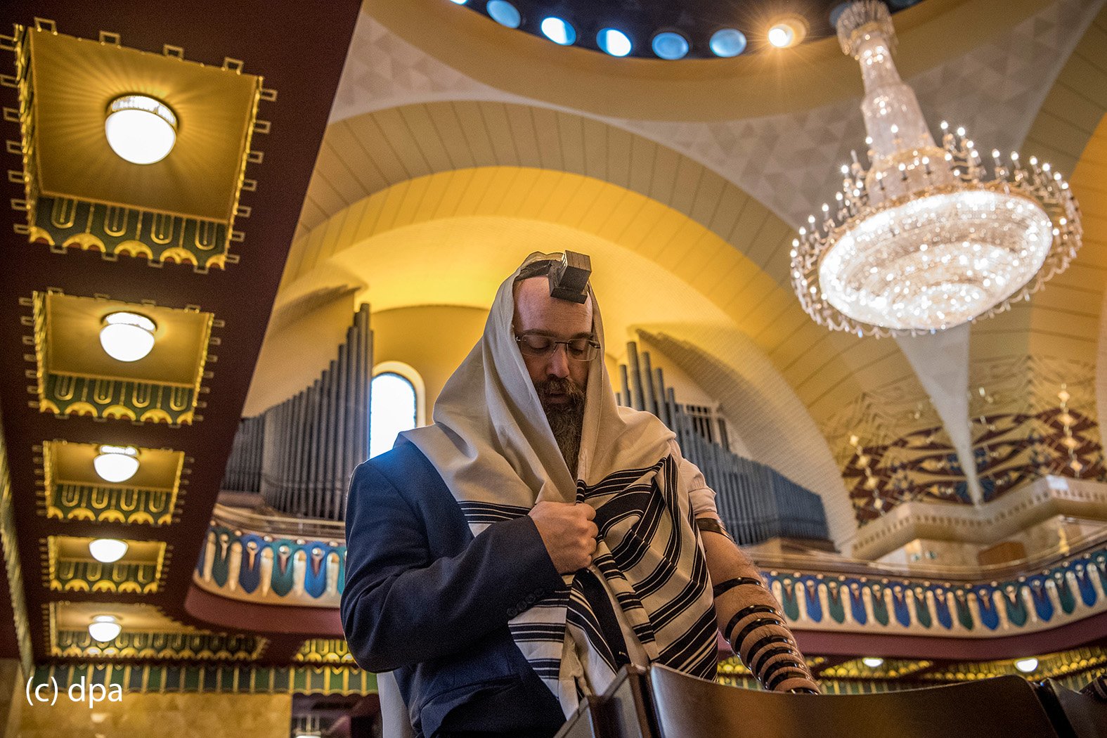 Während sei linker Arm mit einem Gebetsriemen aus Leder umwickelt ist, steht ein Betender in der Westend-Synagoge in Frankfurt am Main in sich versunken unter dem riesigen Leuchter in der Mitte des Gebetssaals. In der Synagoge kommen die Gläubigen aus unterschiedlichen Glaubensausrichtungen bis zu dreimal täglich zum Gebet zusammen. 